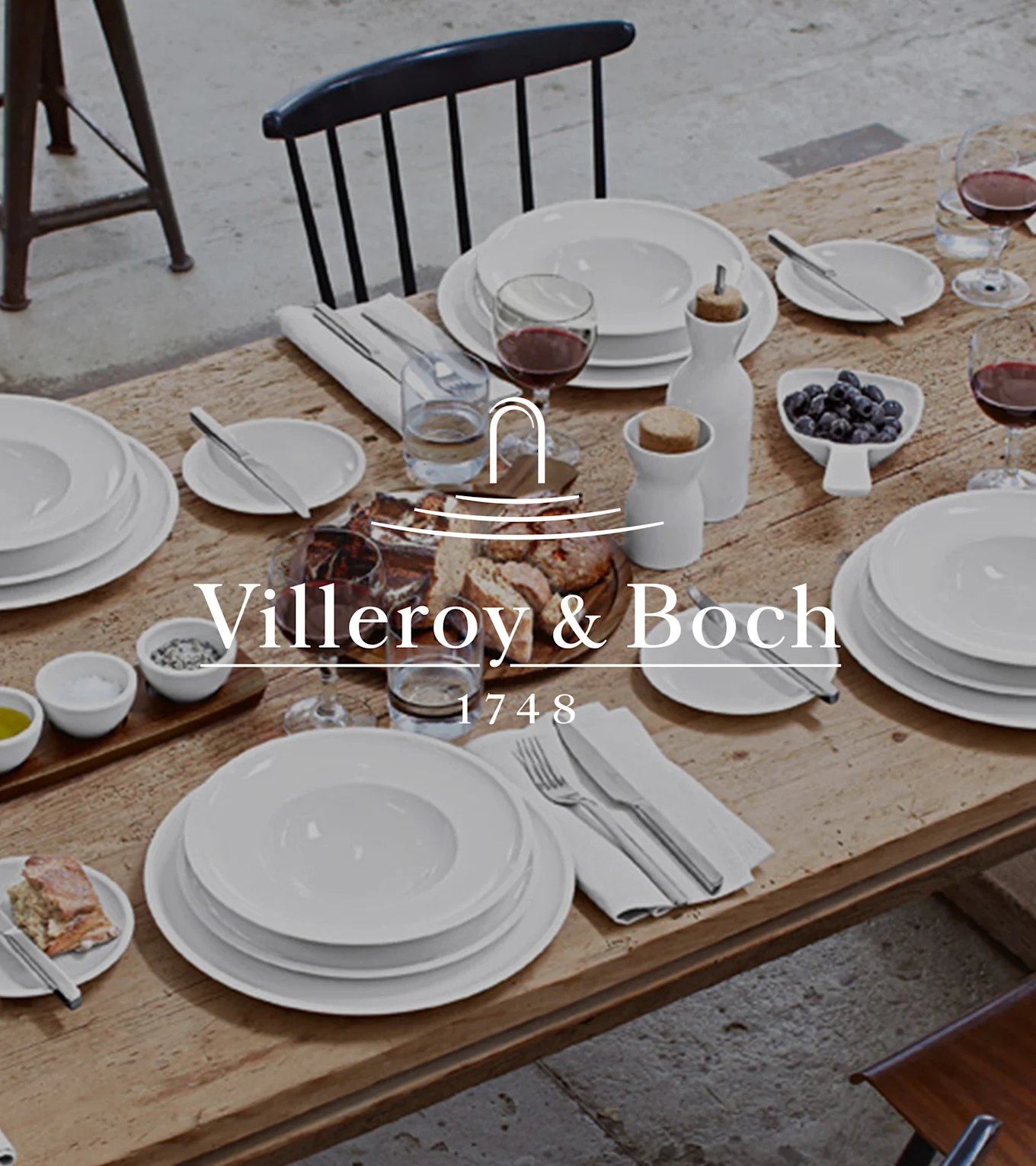 Et rustikt trebord dekket med Villeroy & Boch hvitt servise, vin, brød, blåbær, salt, pepper og olivenolje for et måltid.