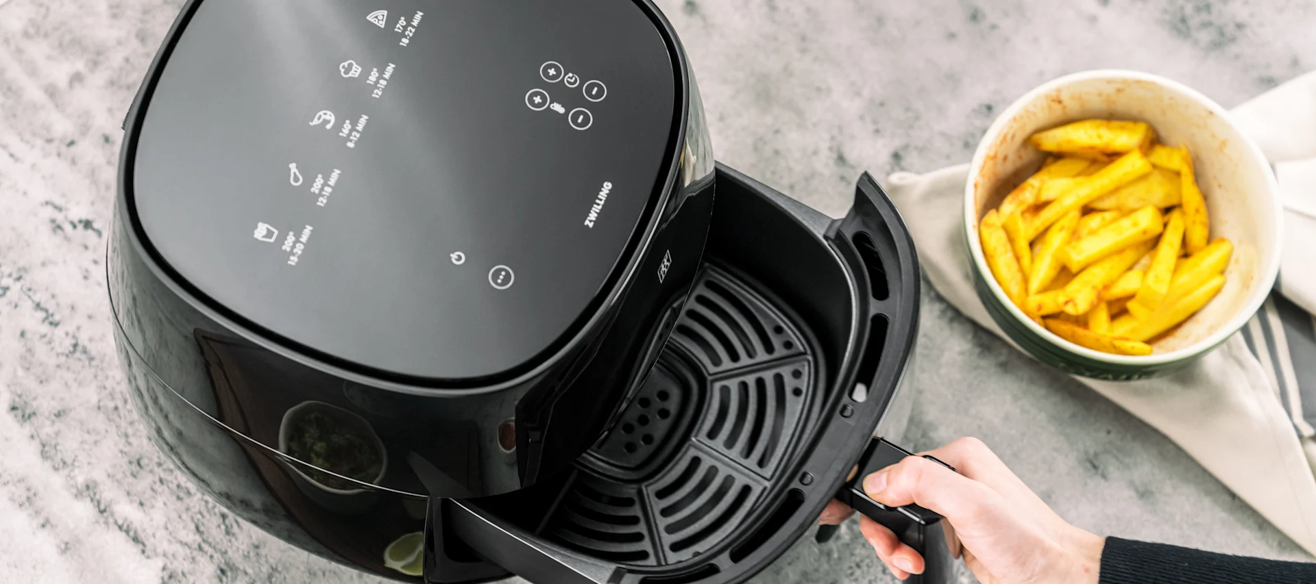 Ovenfra bilde av en hånd som trekker ut kurven fra en svart airfryer, ved siden av en skål med pommes frites.