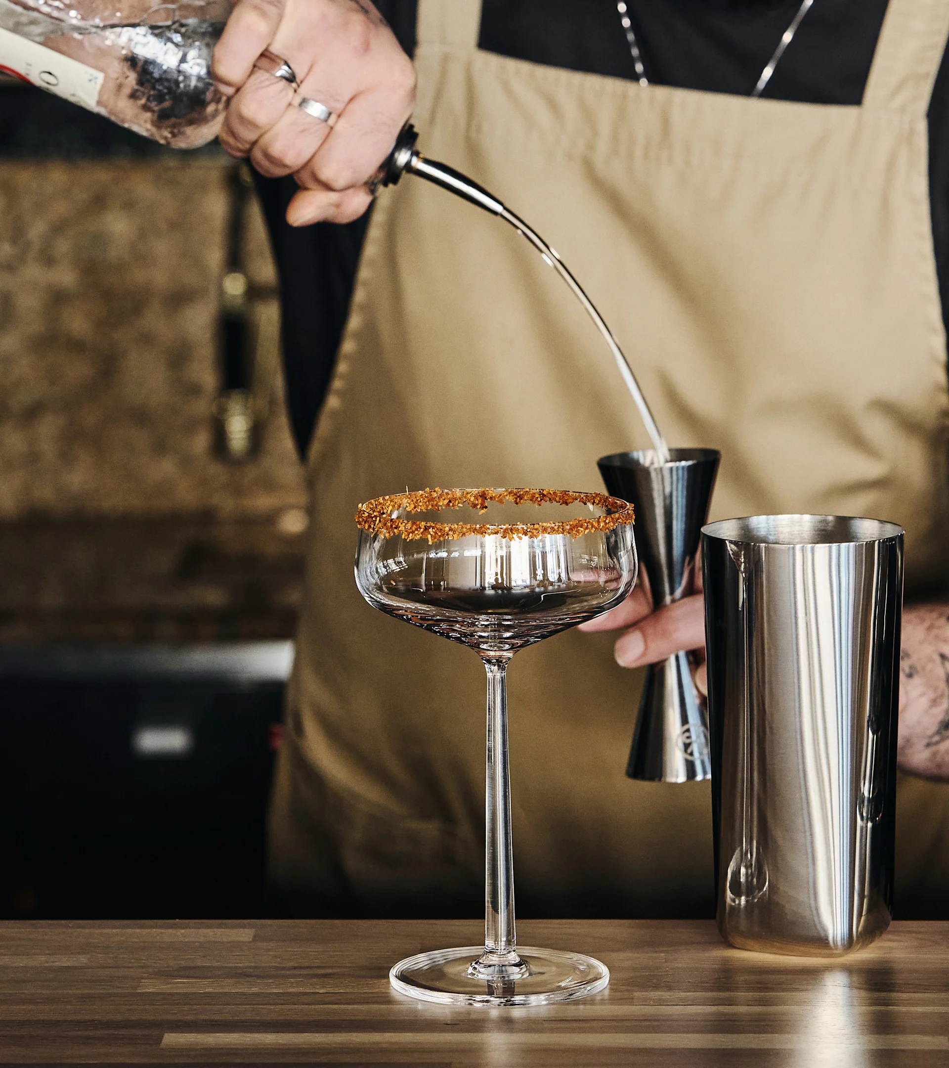 En bartender heller en drink fra en flaske i et måleglass, med et sukkerkantet cocktailglass og en shaker på en trebenk.