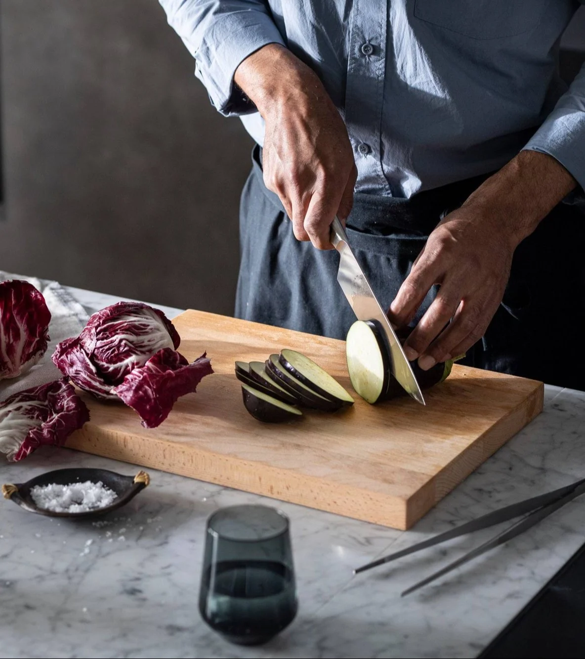 Nærbilde av en person som skjærer aubergine på et trefjøl med radicchio og salt i nærheten.