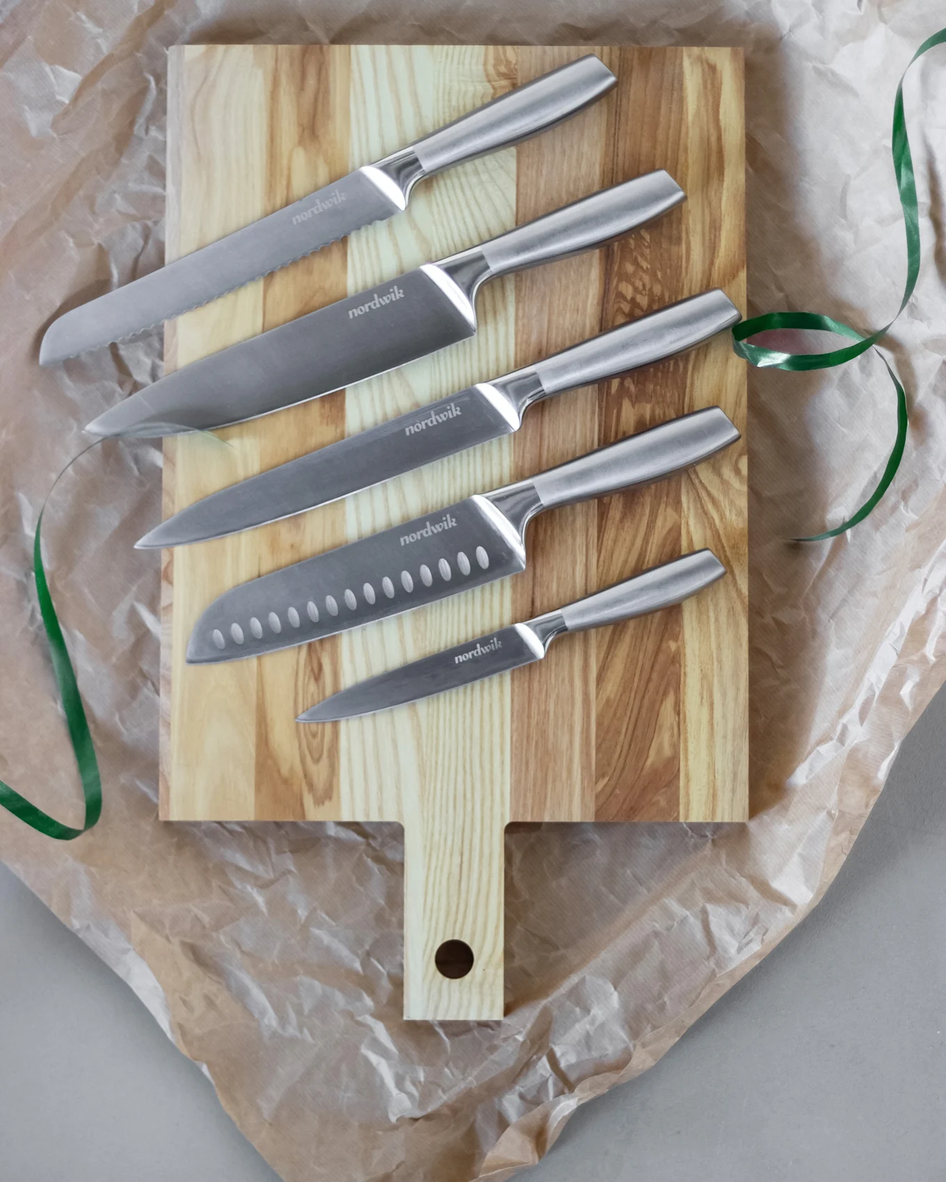 Sett med fem Nordwik kniver i rustfritt stål på et skjærebrett av tre, omgitt av krøllet papir og et grønt bånd.