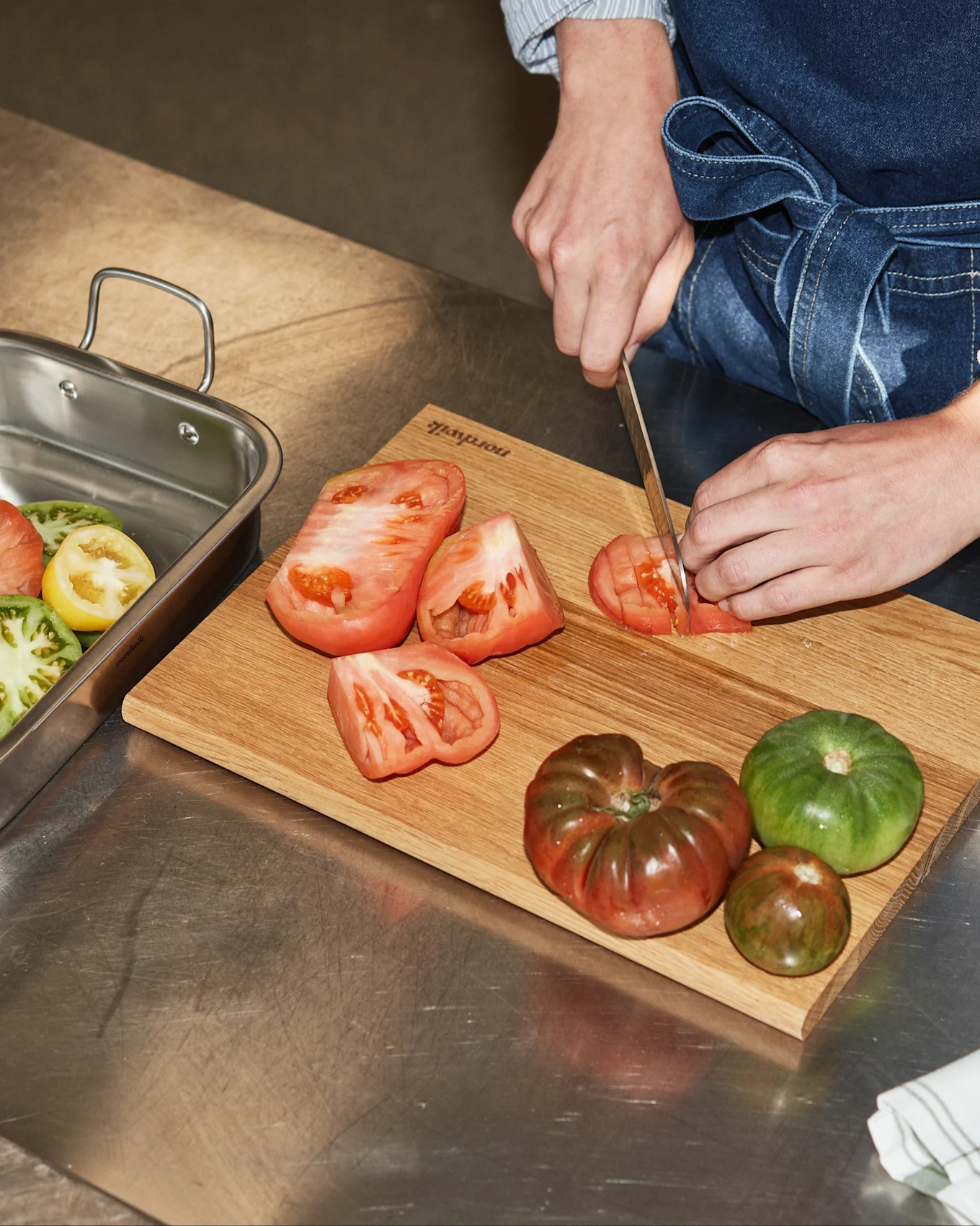 Hender skjærer en rød arvetomat på et skjærebrett av tre, ved siden av en metallform med røde, gule og grønne tomatskiver.