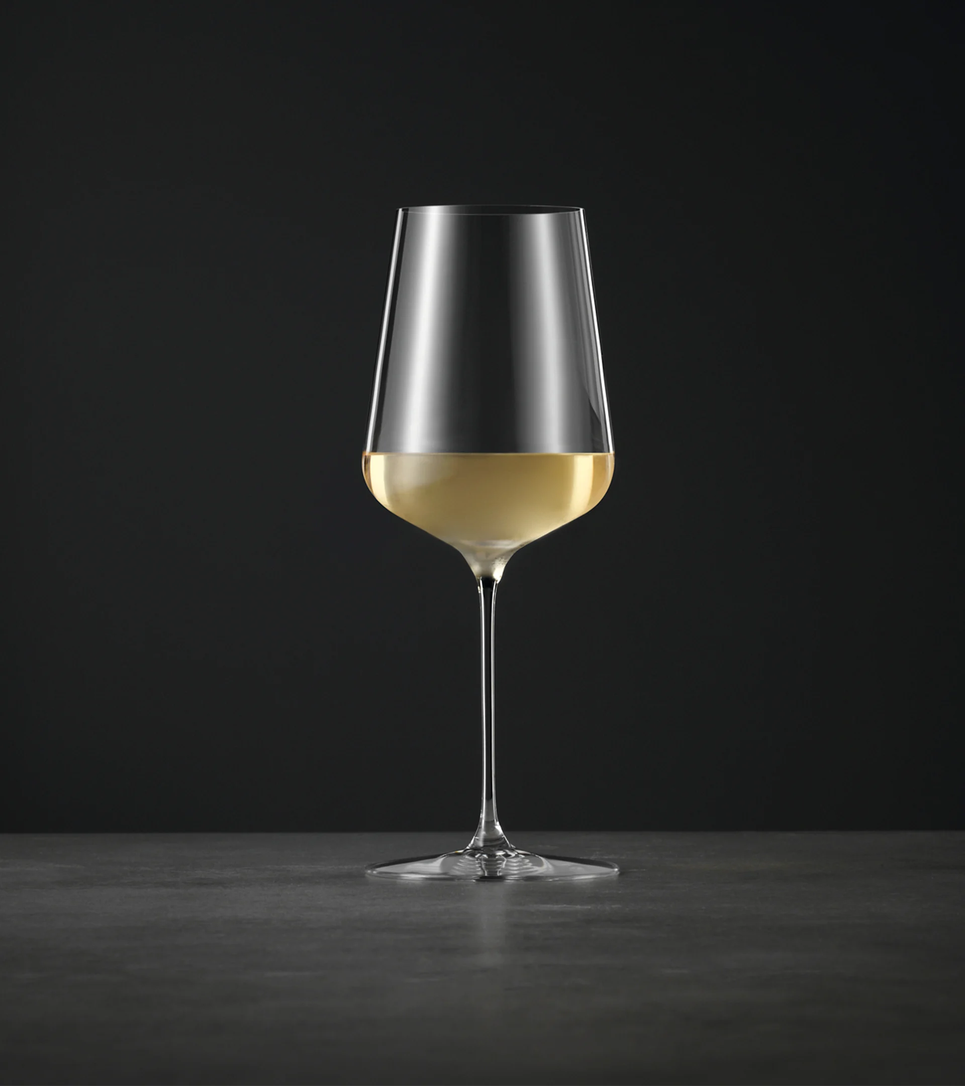Et høyt, elegant vinglass med lang stett, halvveis fylt med lys hvitvin, mot en mørk bakgrunn.
