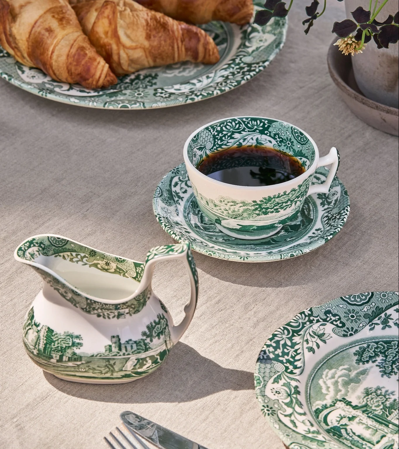 Frokostbord med grønn-hvitt mønstret porselenskopp med kaffe, fløtemugge og tallerkener med croissanter.