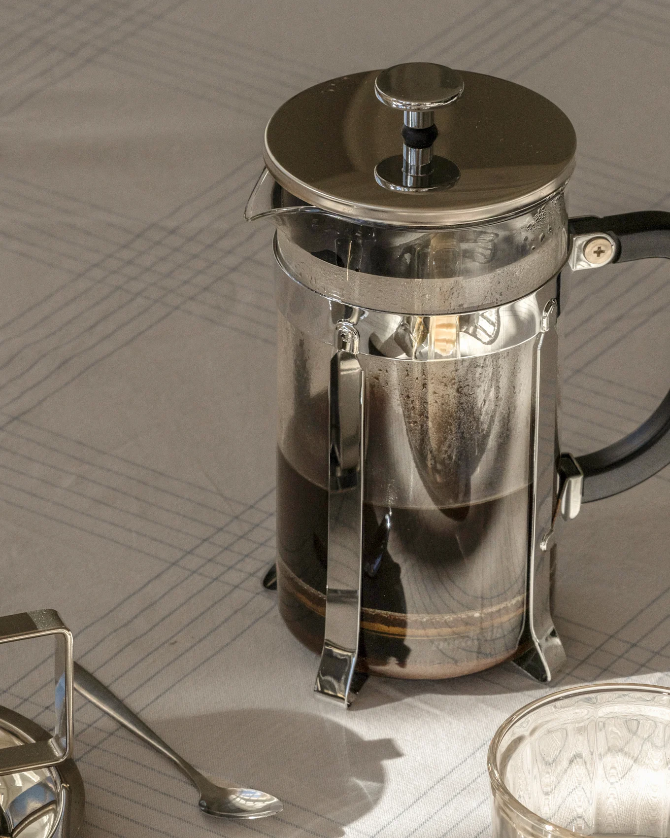 French press kaffekanne med mørk kaffe på en stripet duk, ved siden av en skje og et glass.