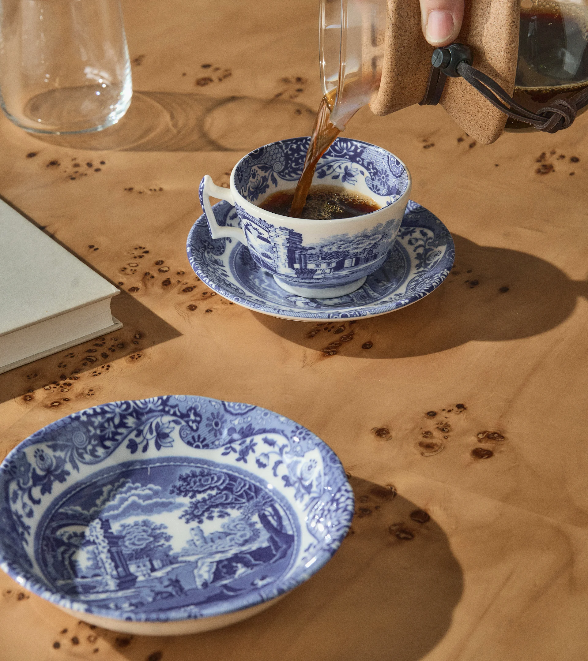 En hånd heller kaffe i en blå og hvit mønstret tekopp på et trebord med en matchende bolle, en bok og et glass.