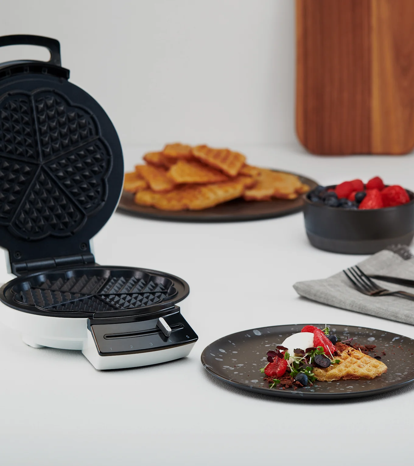 Åpent vaffeljern ved siden av tallerken med vaffel, krem, bær og mikrogrønt. Flere vafler og bær i bakgrunnen.