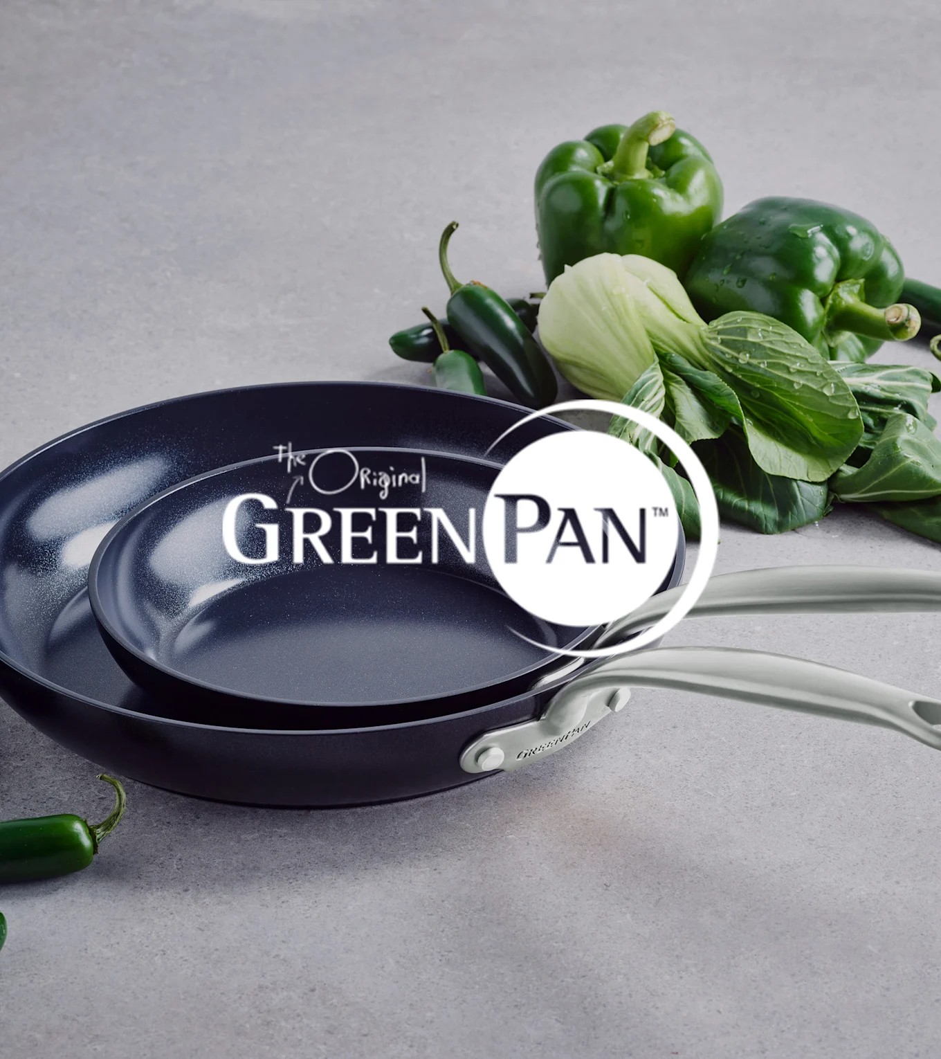 To mørke non-stick panner med lyse håndtak, stablet, ved siden av grønn paprika, pak choi og jalapeños. GreenPan logo.