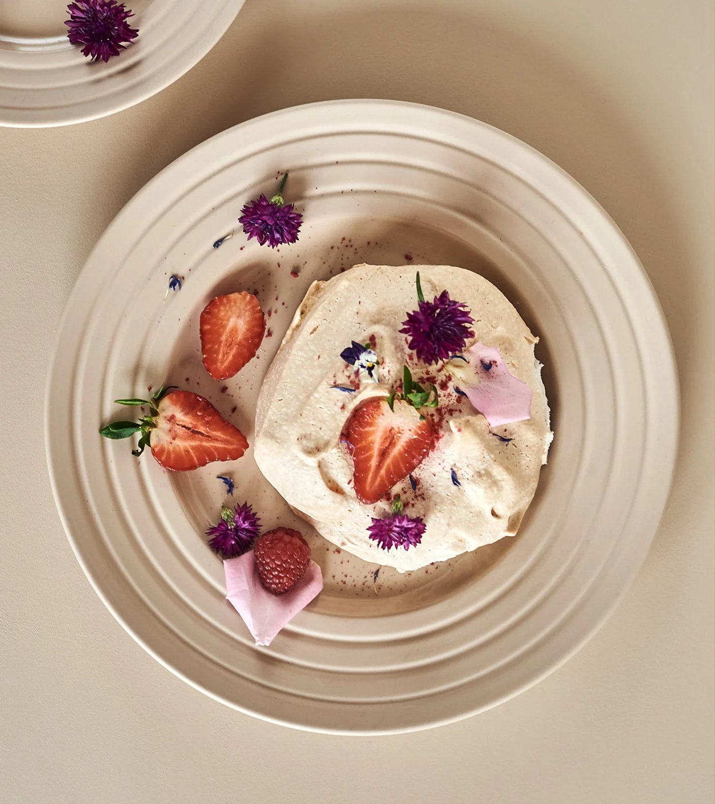 En kremet dessert med jordbær, et bringebær og lilla blomster på en beige riflet tallerken.
