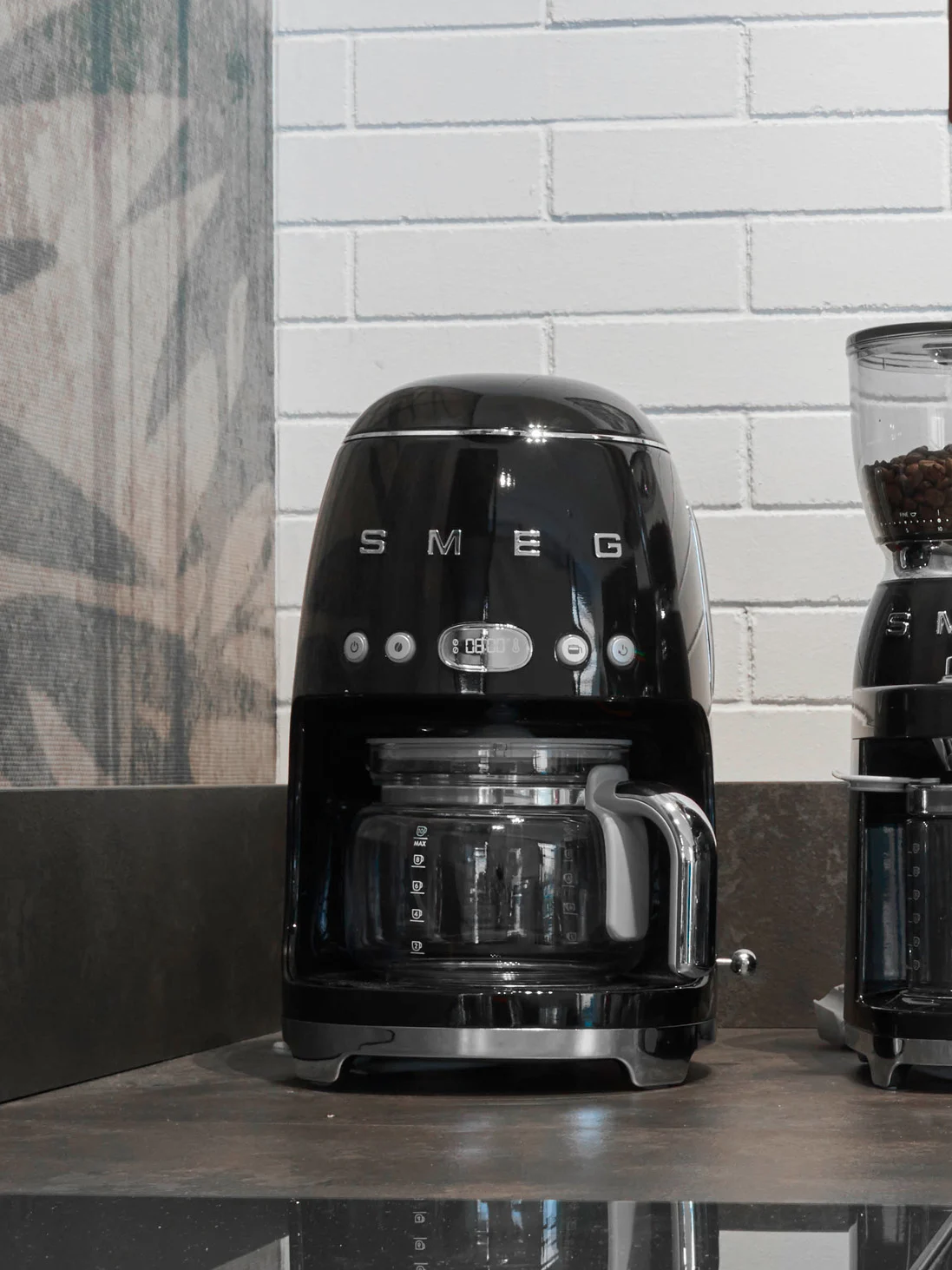 Svart Smeg kaffemaskin med glasskanne og digitalt display som viser '08:00' på en mørk benkeplate.