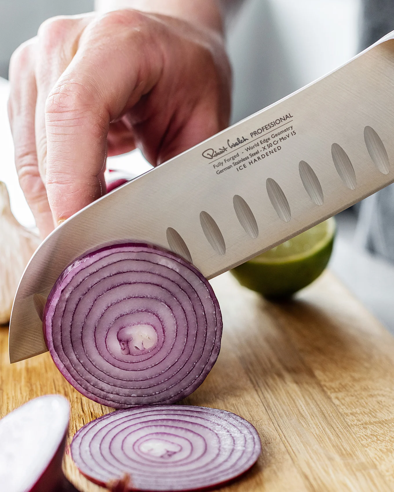 En kokkekniv med teksturert blad skjærer en rødløk på et skjærebrett av tre, med en lime i bakgrunnen.