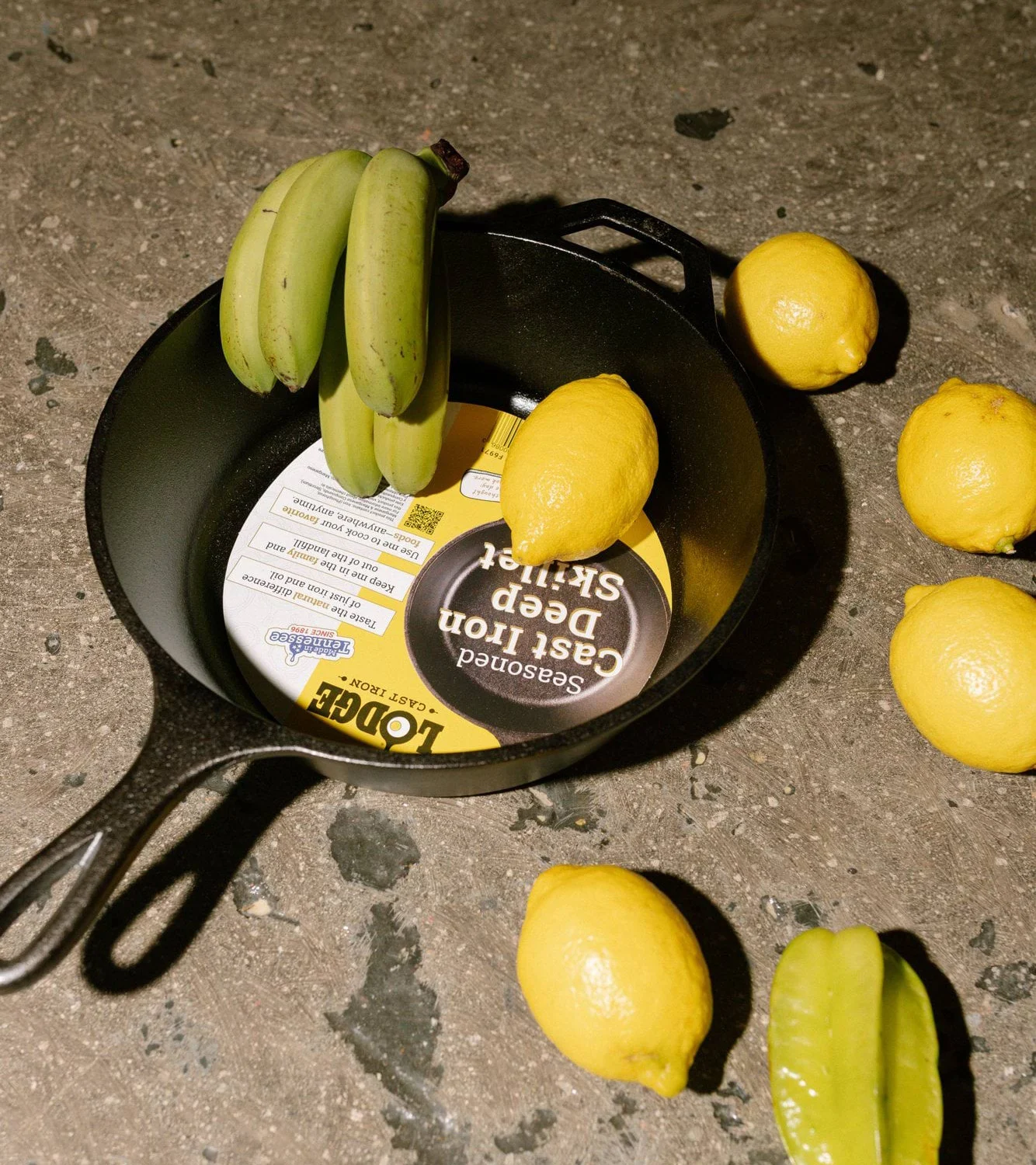 Støpejernspanne med etikett inni, umodne bananer, sitroner og en stjernefrukt på en mørk overflate.