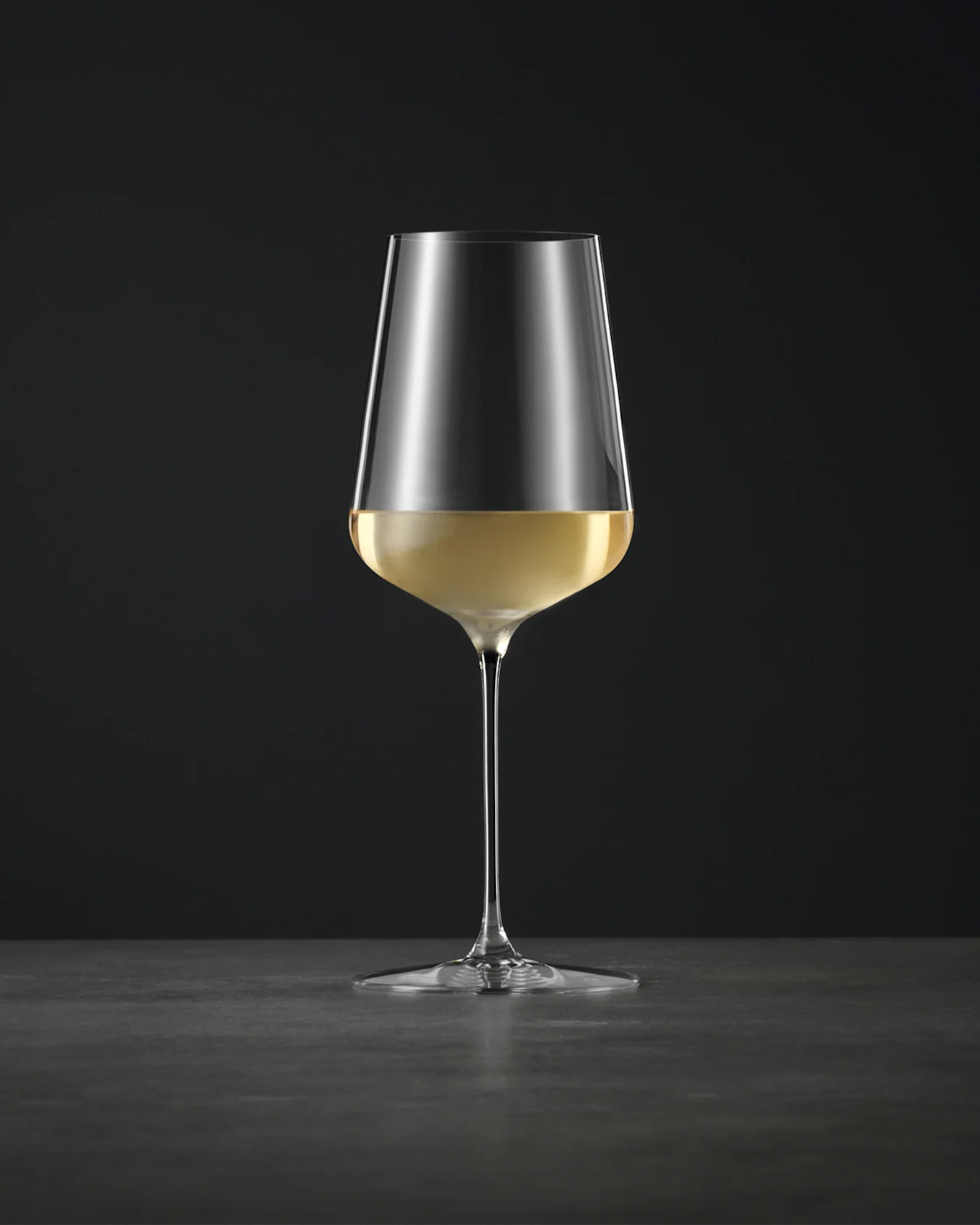 Et enkelt, elegant vinglass fylt med hvitvin, på en mørk overflate mot en svart bakgrunn.