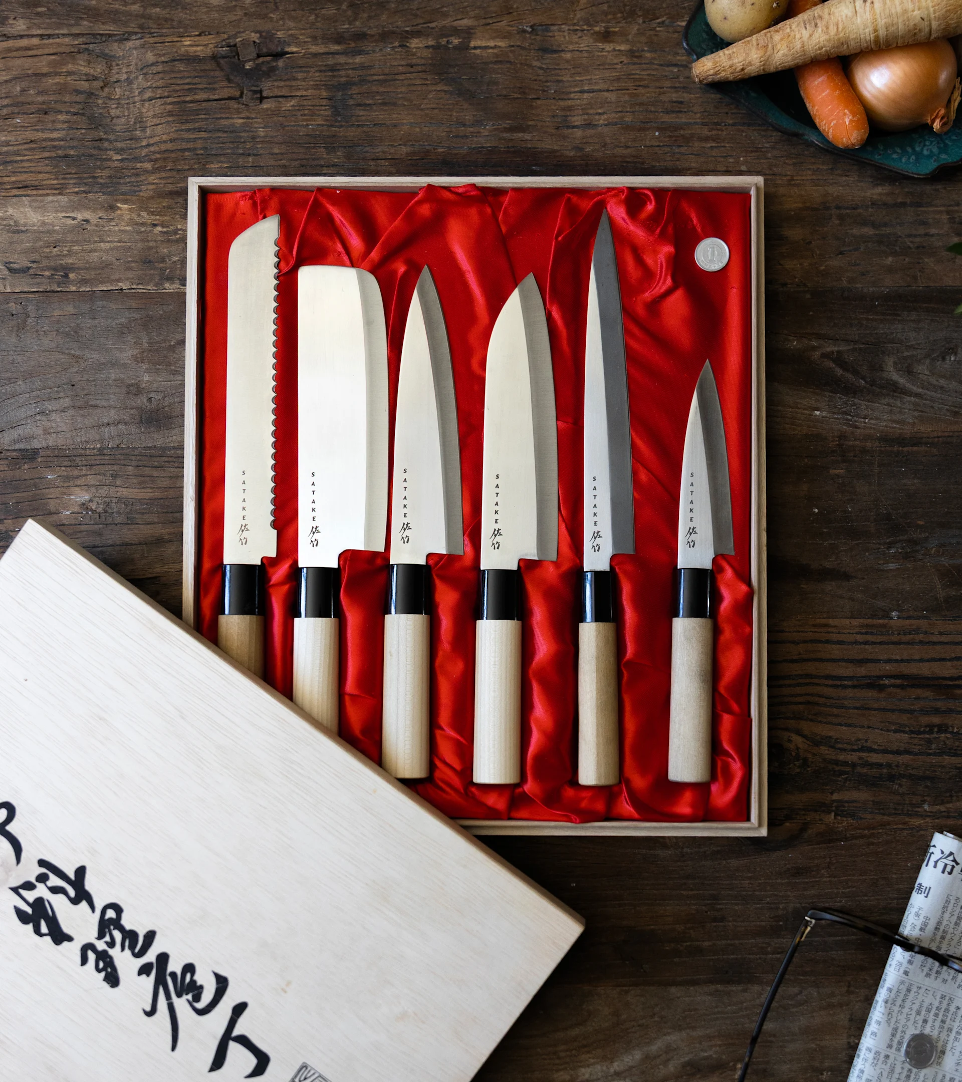 Seks japanske kniver med trehåndtak i en rødforet eske på et trebord, med grønnsaker og avis i bakgrunnen.