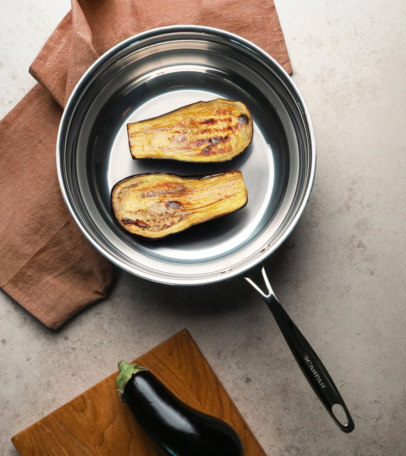 To grillede auberginehalvdeler i en stekepanne av rustfritt stål, med en rå aubergine på et skjærebrett i nærheten.