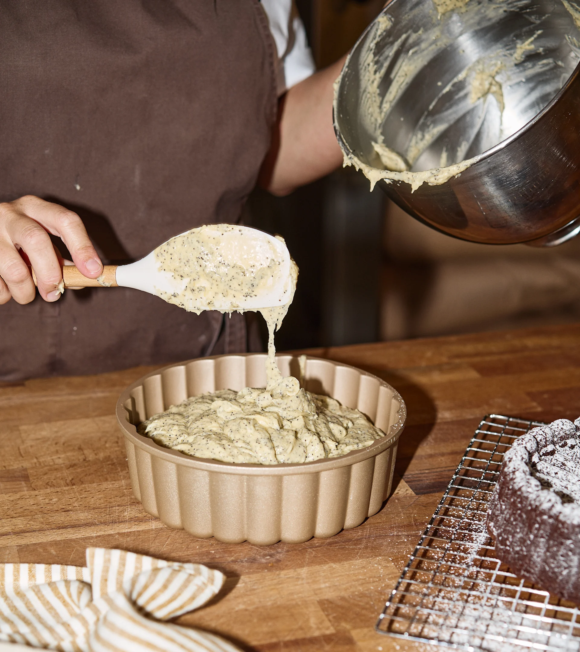 Person øser valmuefrødeig i en gullfarget riflet bakeform. En sjokoladekake med melis er i bakgrunnen.