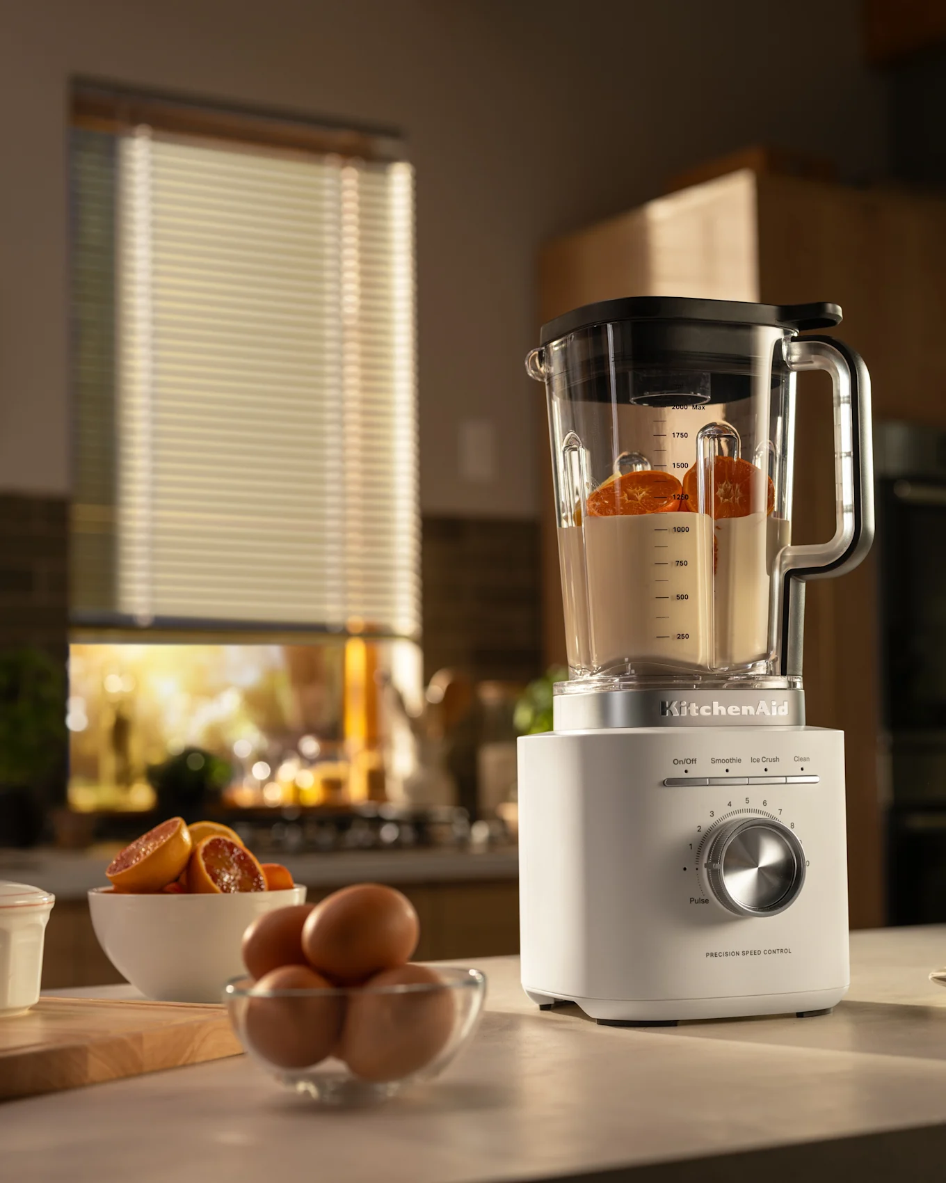 Hvit KitchenAid blender med appelsinskiver og væske, sammen med egg, mel og appelsinkake i et solrikt kjøkken.