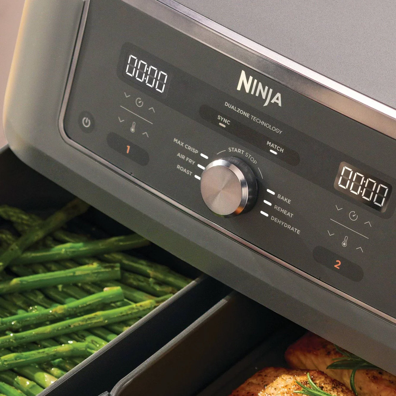 Et Ninja airfryer-kontrollpanel med to digitale skjermer og en knott, som viser grønn asparges i den ene skuffen og kylling i den andre.