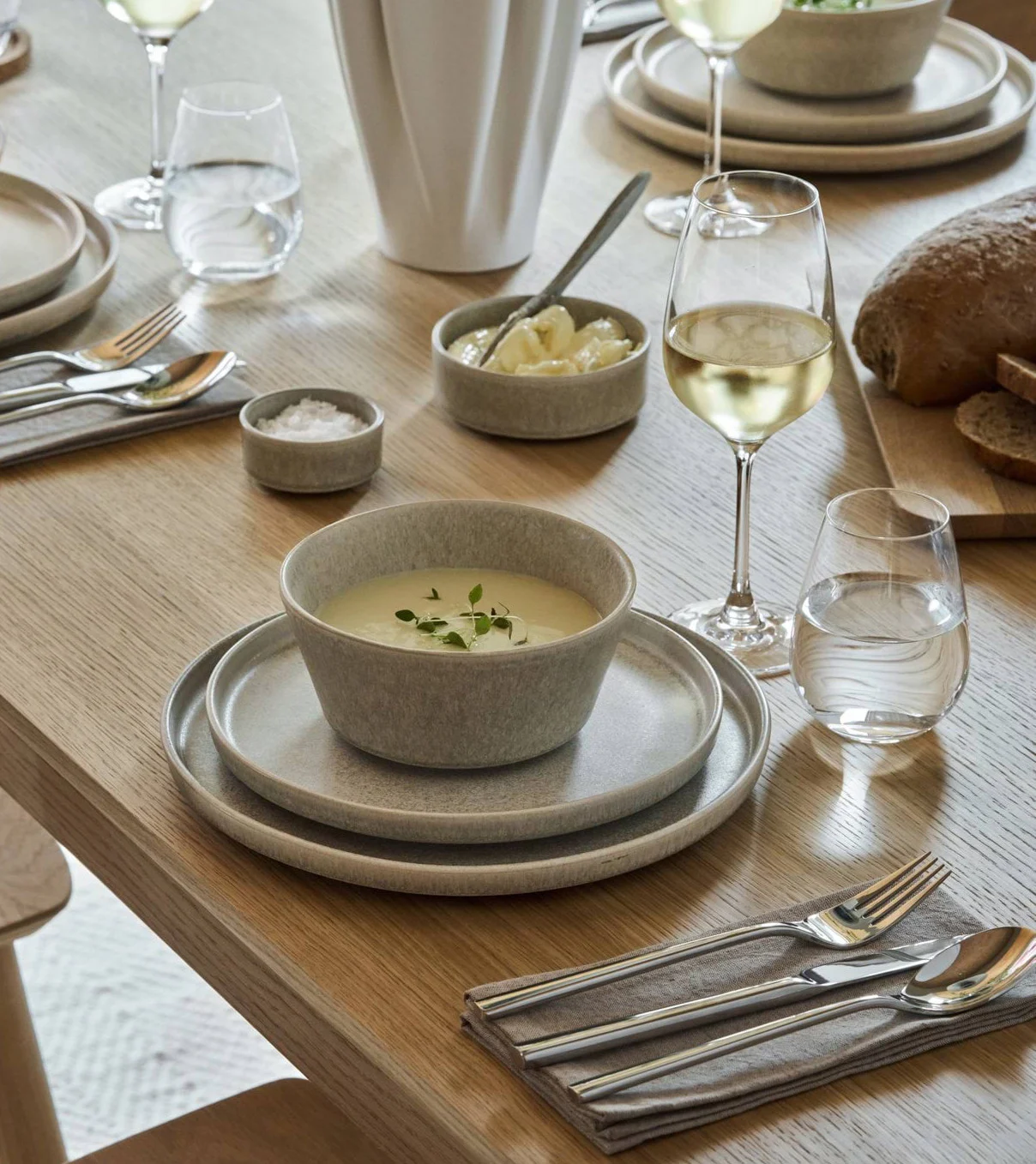 Et spisebord dekket med lysegrått steintøy, suppe, brød, vin, vann og bestikk.