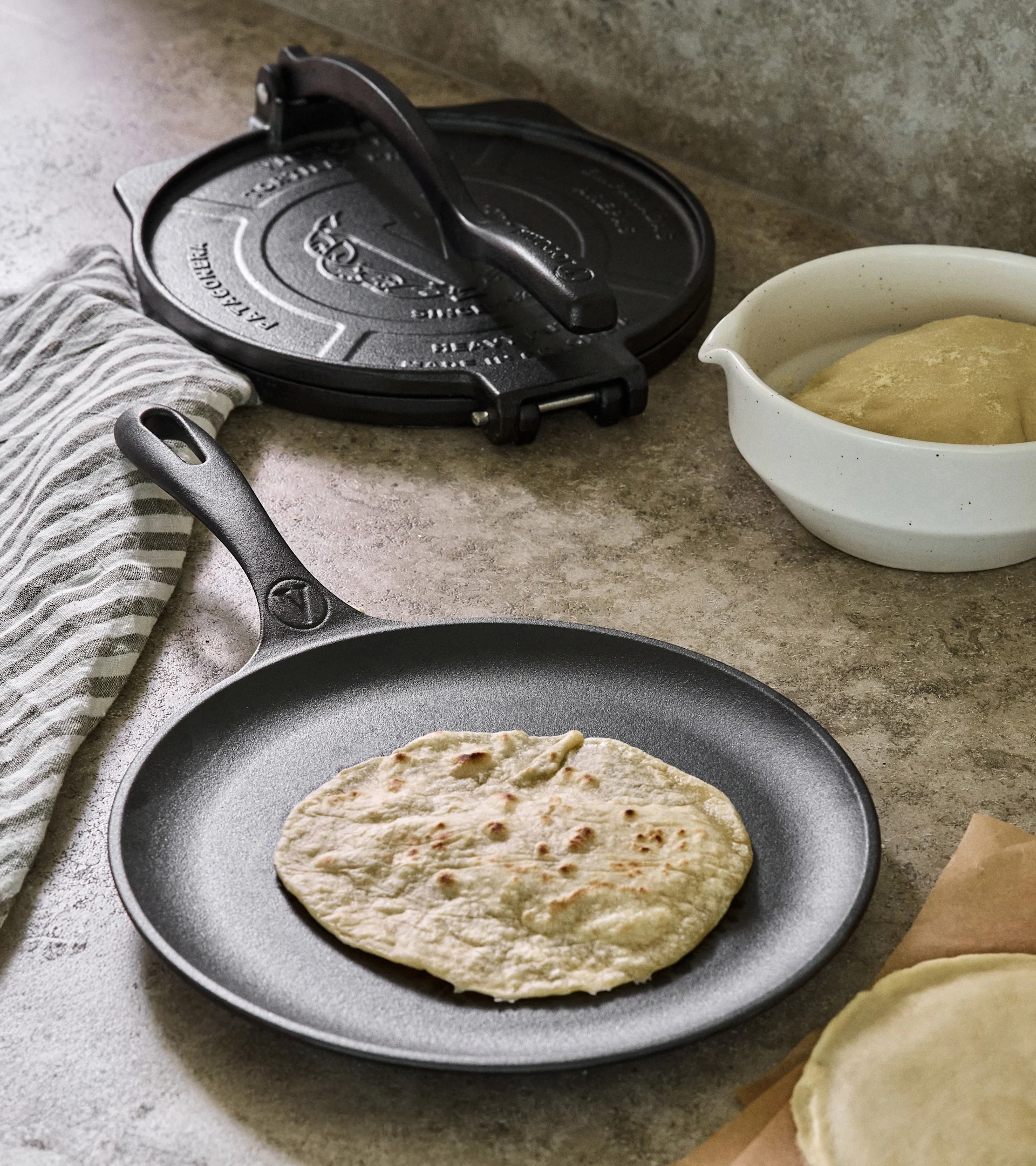 Tortillapresse og stekepanne i støpejern med stekt tortilla, deig og rå tortillas på en benkeplate.