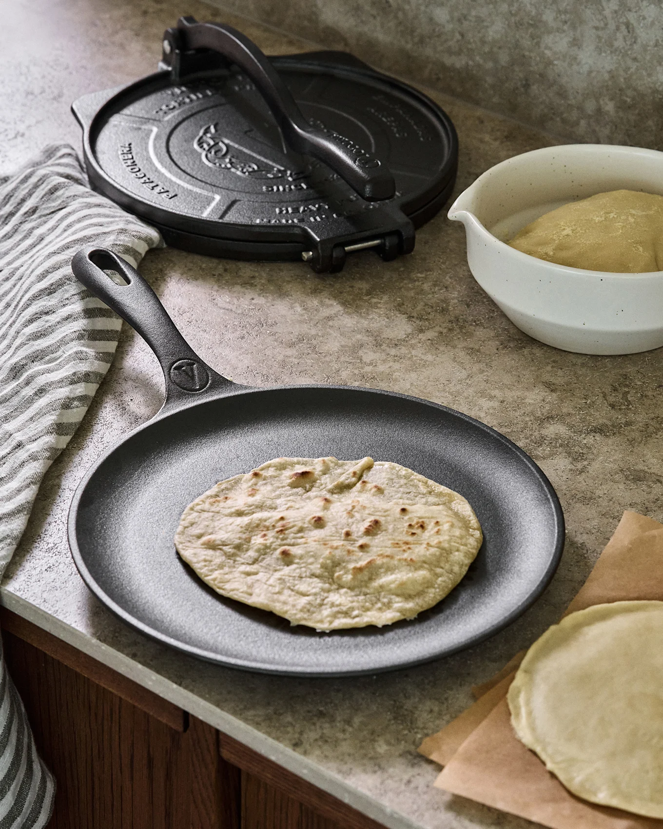 Støpejernspanne med flatbrød, ved siden av en tortillapresse og deig.