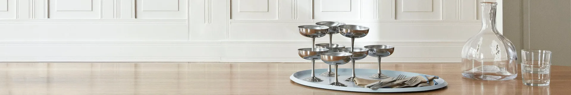 Et lyseblått ovalt brett med sølv champagneglass, bestikk og serviett på et trebord ved siden av en glasskaraffel.