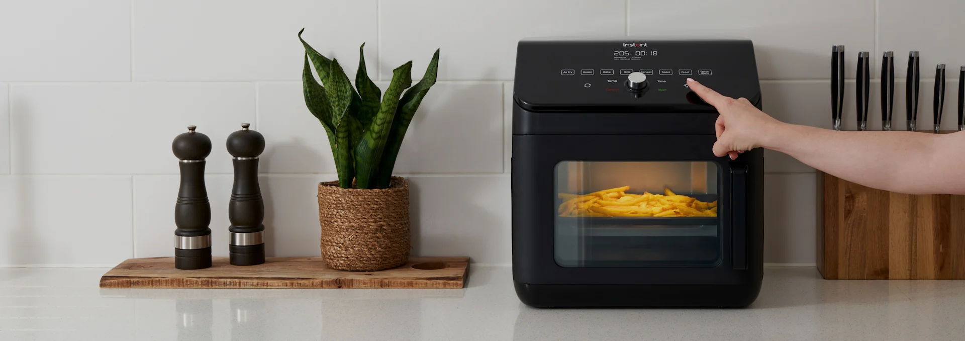 Hånd trykker på knapp på sort airfryer med pommes frites, ved siden av krydderkverner, en plante og knivblokk på benken.