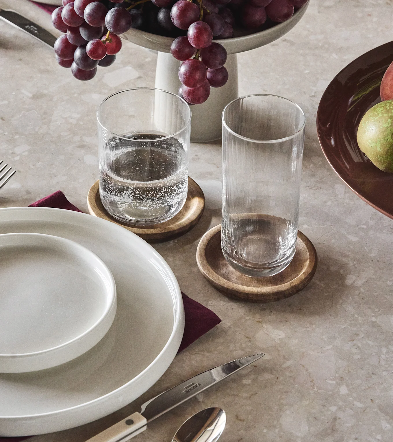 Spisebord med hvite tallerkener, to klare glass på treunderlag, røde druer og et stykke frukt.