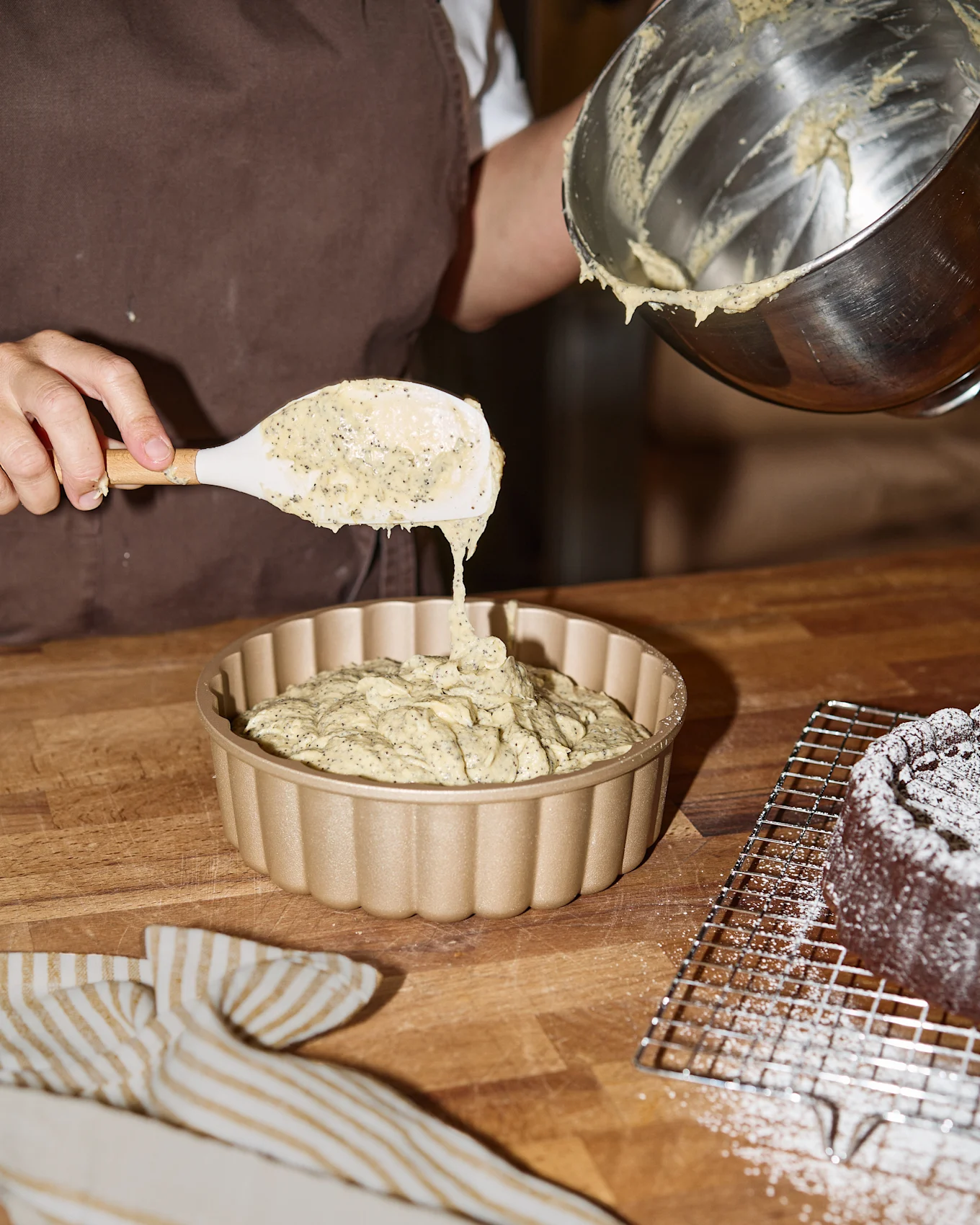Hender skjeer valmuefrødeig i en gullriflet kakeform, med en bakt, melisdryss-kake i nærheten.