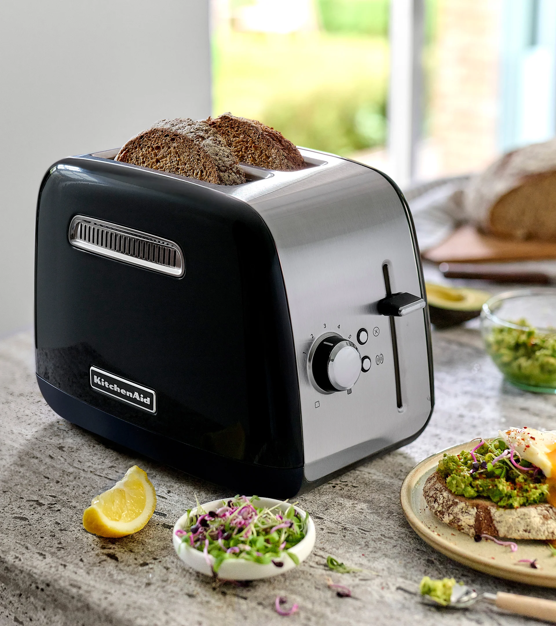 En svart og rustfri KitchenAid brødrister med to skiver mørkt brød. På benken, avokadotoast med egg, spirer og en sitronbåt.