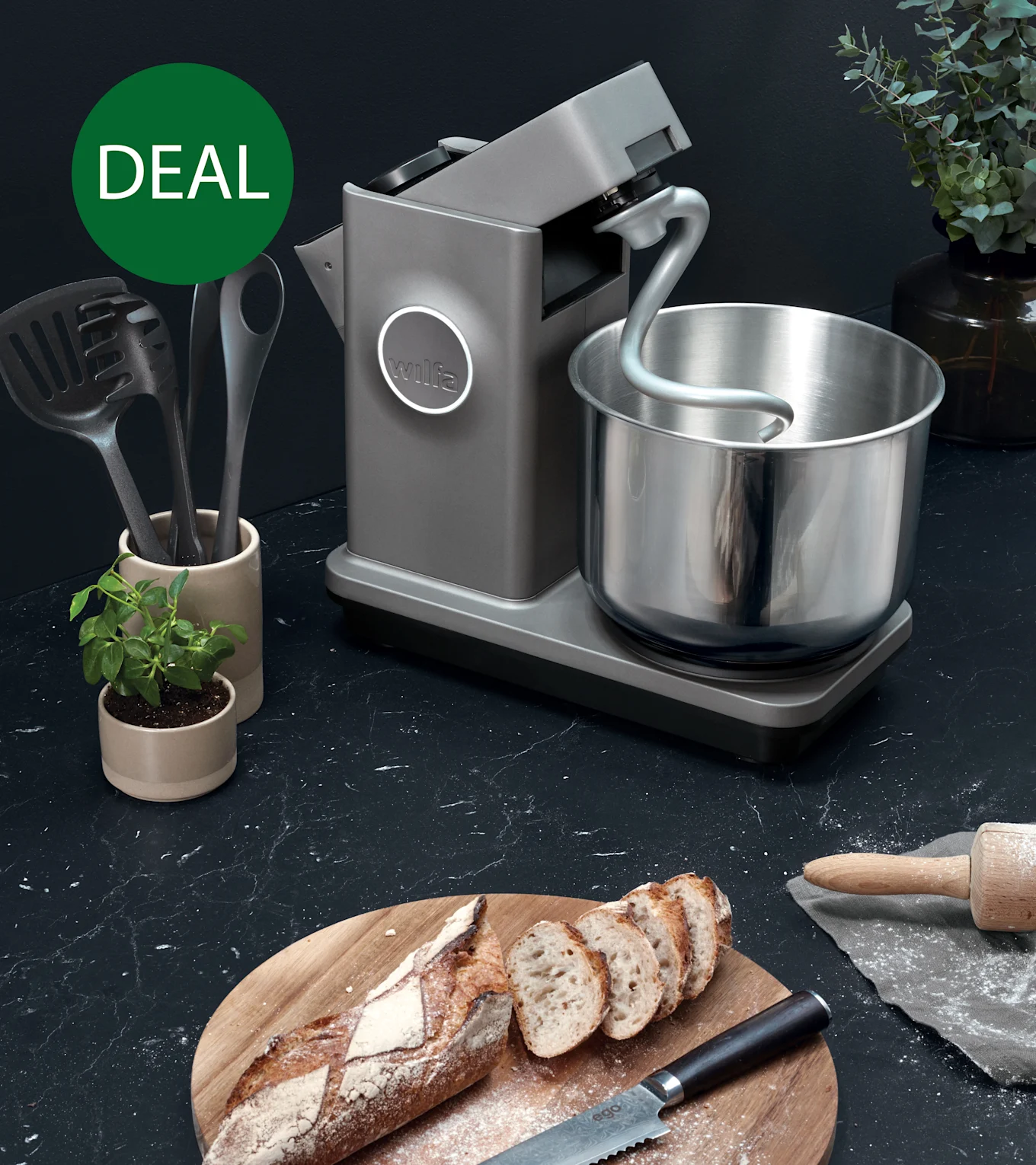 Grå kjøkkenmaskin med eltekrok og stålbolle på mørk benkeplate med skåret brød, markert med grønn 'TILBUD'-sirkel.