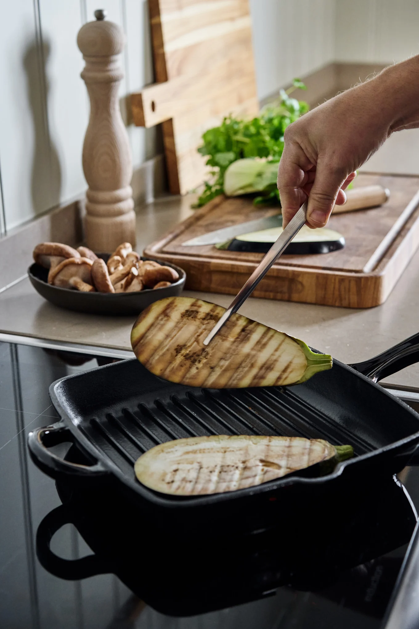 Hånd snur grillede aubergineskiver i svart støpejernsgrillpanne på koketopp. Ingredienser er i bakgrunnen.