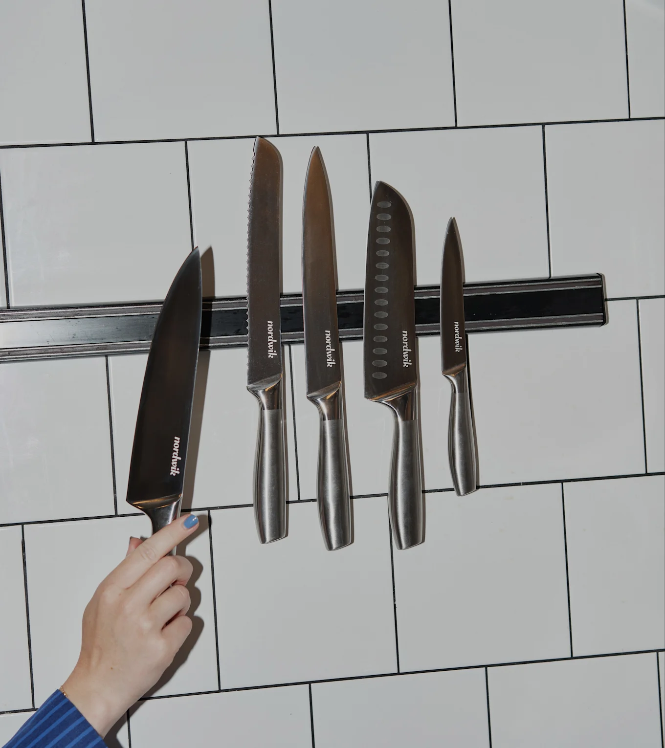 En hånd med blå neglelakk holder en Nordwik kokkekniv nær en magnetlist med fire andre Nordwik kniver på en hvit flislagt vegg.