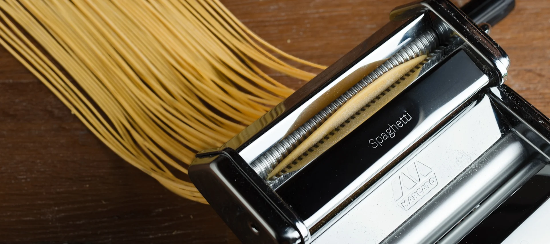 Fersk spaghetti ekstruderes fra en pastamaskin ned på en treflate.
