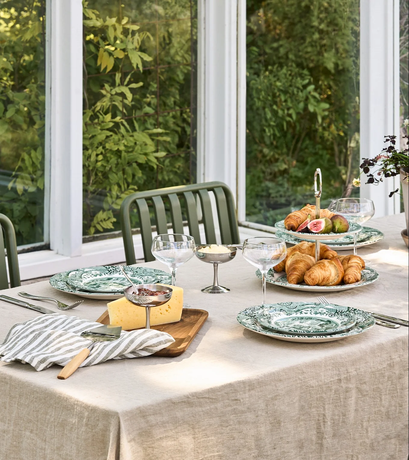 Et frokostbord med grønne mønstrede tallerkener, croissanter, fiken, ost, syltetøy og smør i en lys vinterhage.