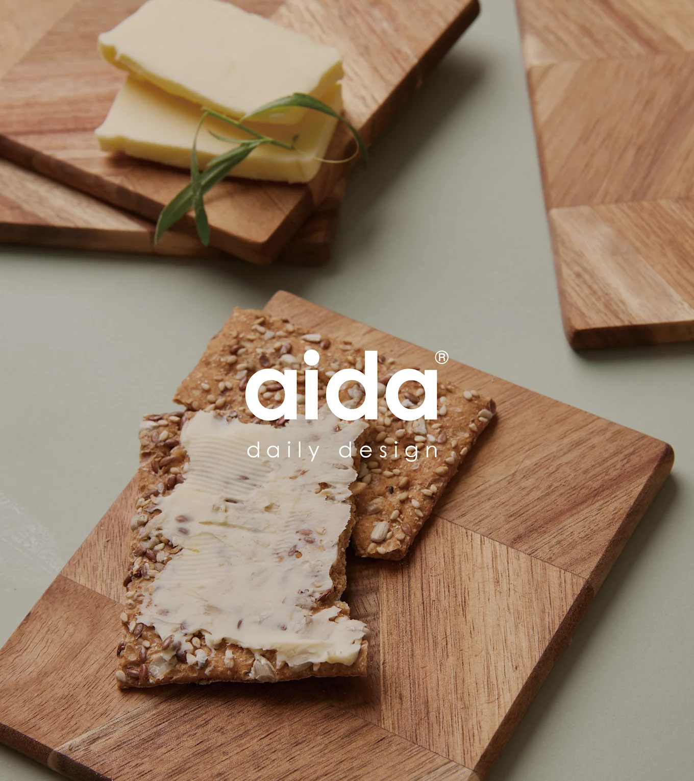 Trebrett med geometriske mønstre. Ett holder en kjeks med smør, det andre smørskiver og en urt. Aida-logo.