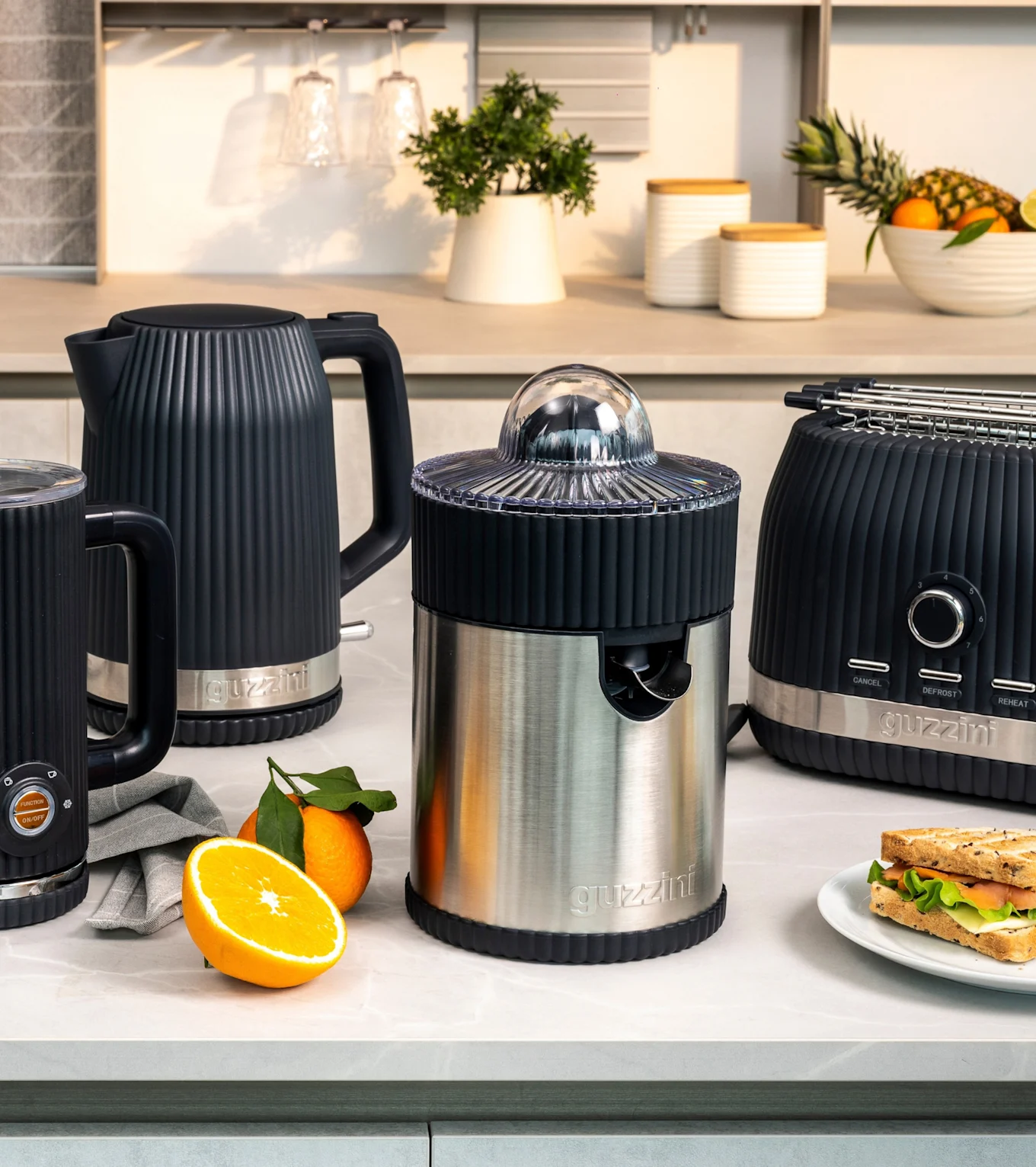 Svart riflet vannkoker, melkeskummer, rustfri ståljuicer og brødrister på kjøkkenbenk med appelsiner og sandwich.