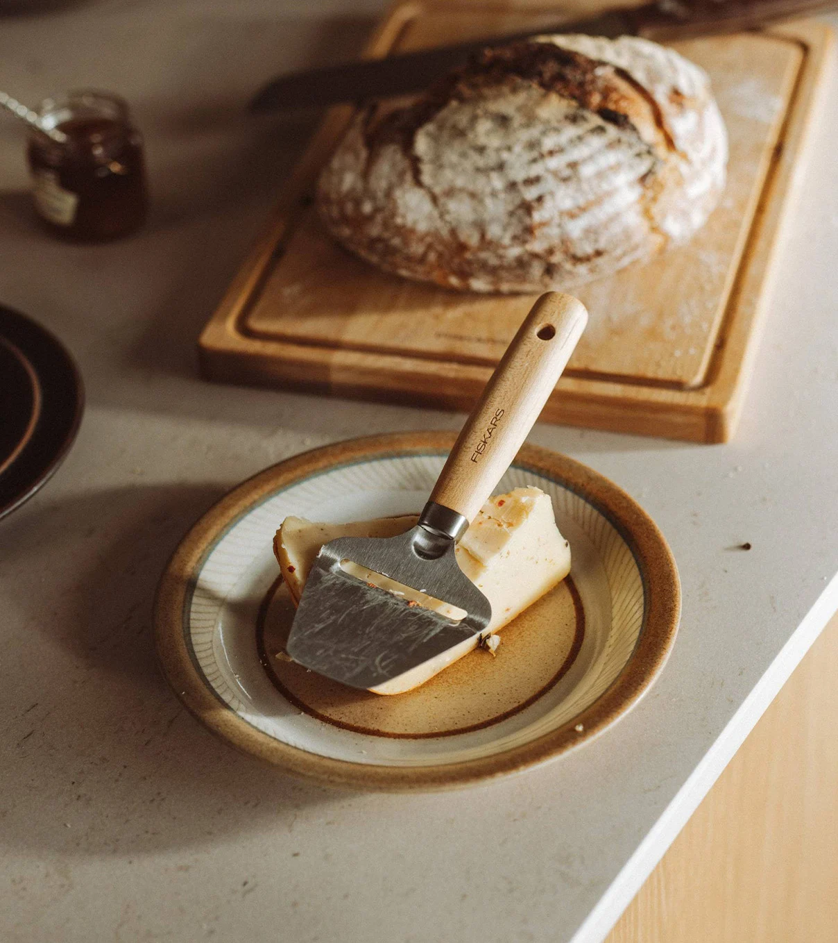 En osteskjærer med trehåndtak hviler på en osteblokk på en tallerken, med et brød i bakgrunnen.