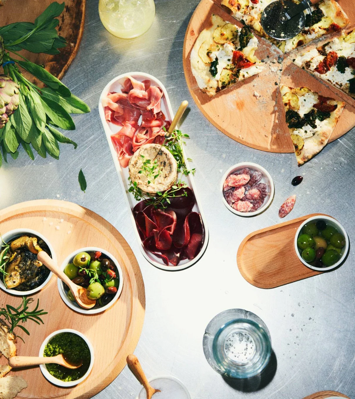 Oversiktsbilde av matbord med skåret pizza, spekematfat, oliven, pesto og tapenade på trefat.