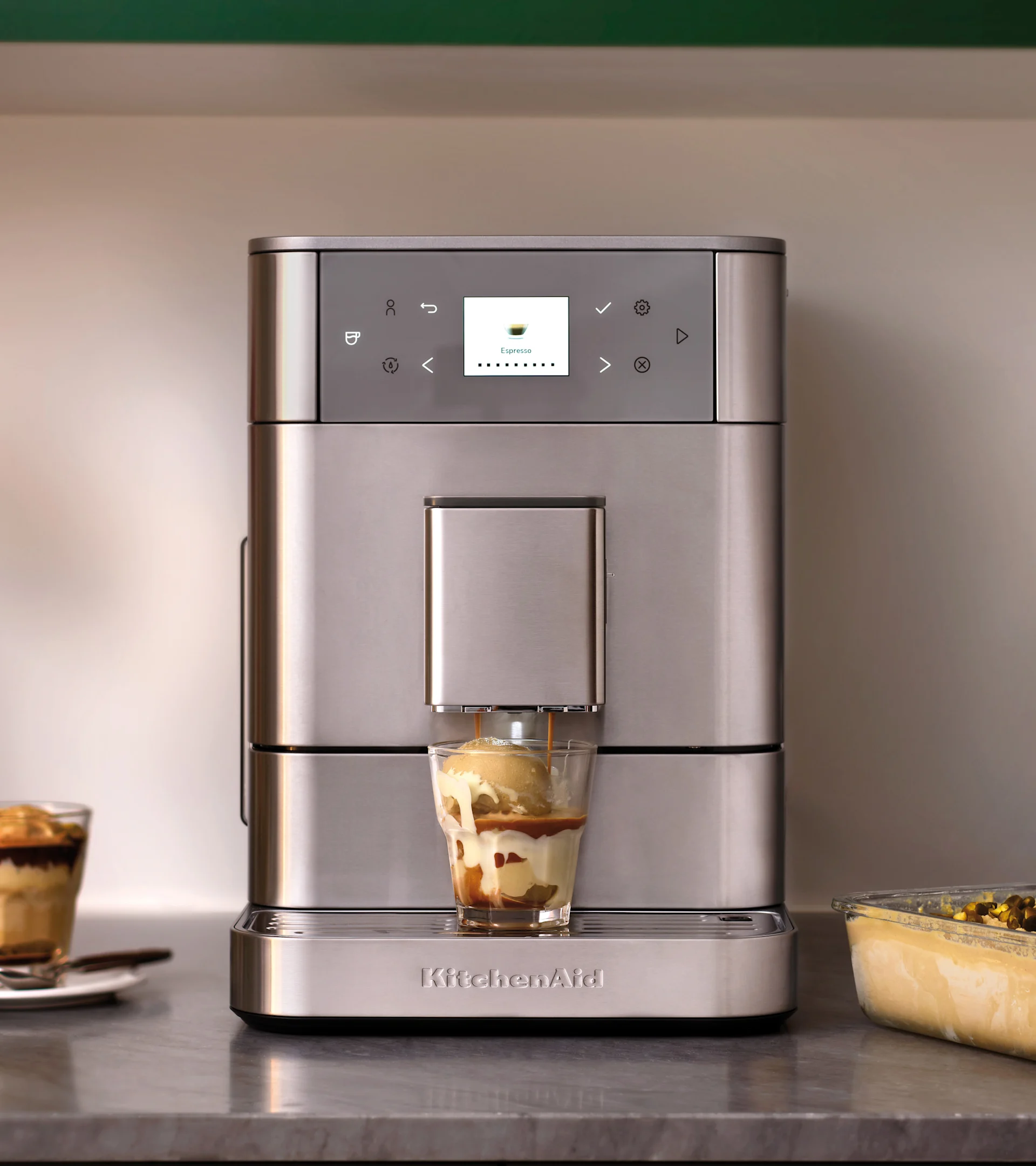 Sølvfarget KitchenAid espressomaskin lager affogato i et glass med is. En annen dessert er i bakgrunnen.