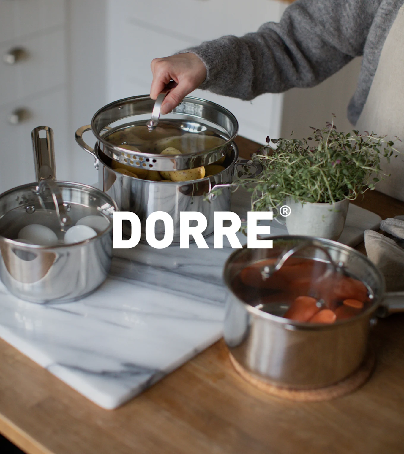 Hånd løfter lokk fra dampkjele i rustfritt stål med poteter, ved siden av kjeler med egg og gulrøtter. DORRE-logo.